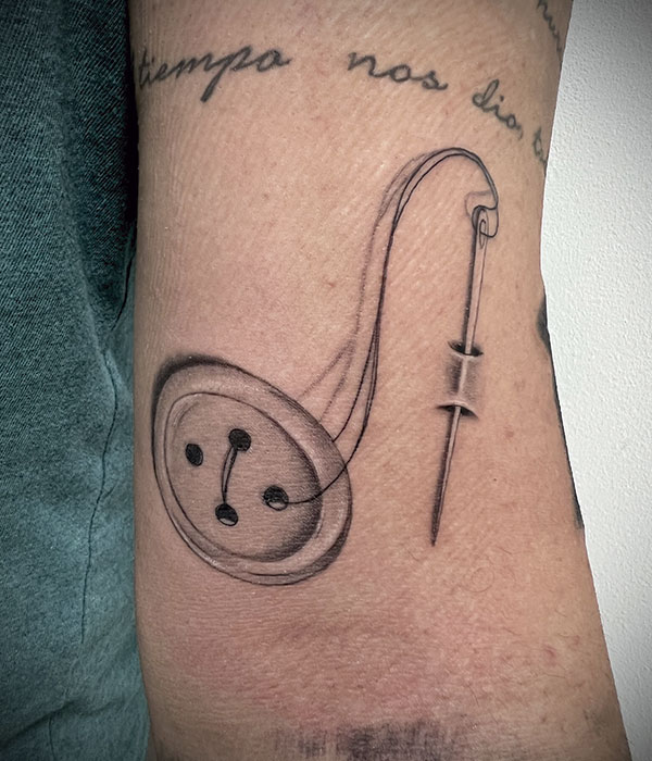 Tatuaje aguja integrada en la piel que acaba de coser un boton, en negro en el brazo. De la Rocha Tattoo Cartagena, Murcia